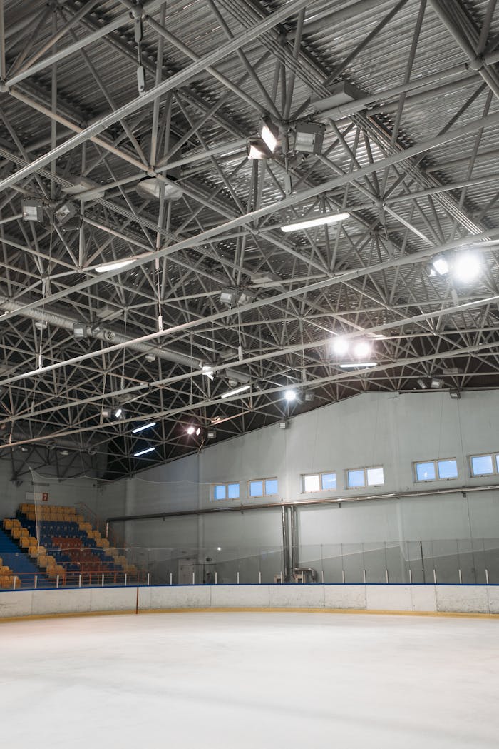 Spacious indoor ice skating rink under a metal truss roof with bright lighting.